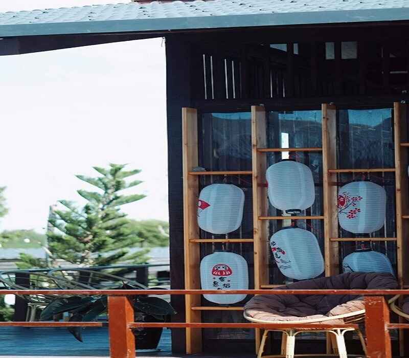 Outdoor seating area with a wooden frame displaying decorative white lanterns, a wicker chair with a cushion, and a view of greenery in the background.