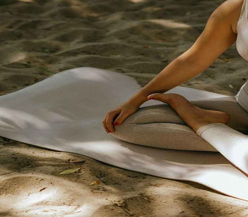 Person meditating outdoors on a yoga mat, sitting cross-legged on sandy ground, with sunlight filtering through trees.