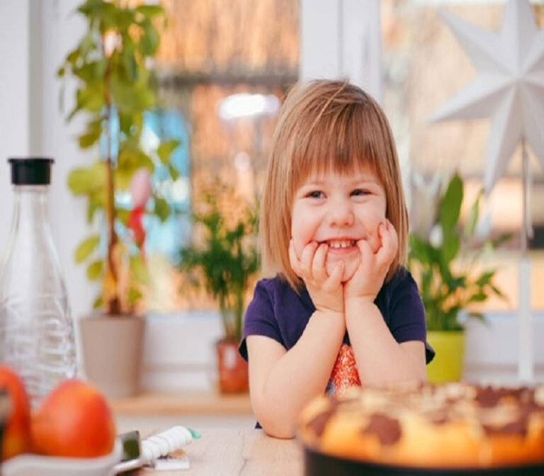 Smiling child sitting at a table with hands on their cheeks, surrounded by a cozy, plant-filled interior.