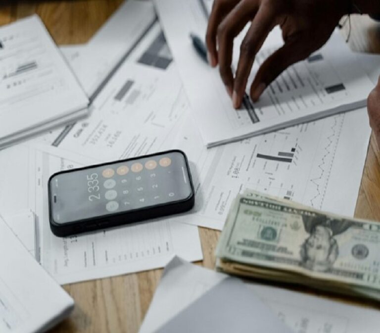 Person calculating finances with a smartphone calculator, documents, and cash on a wooden desk.