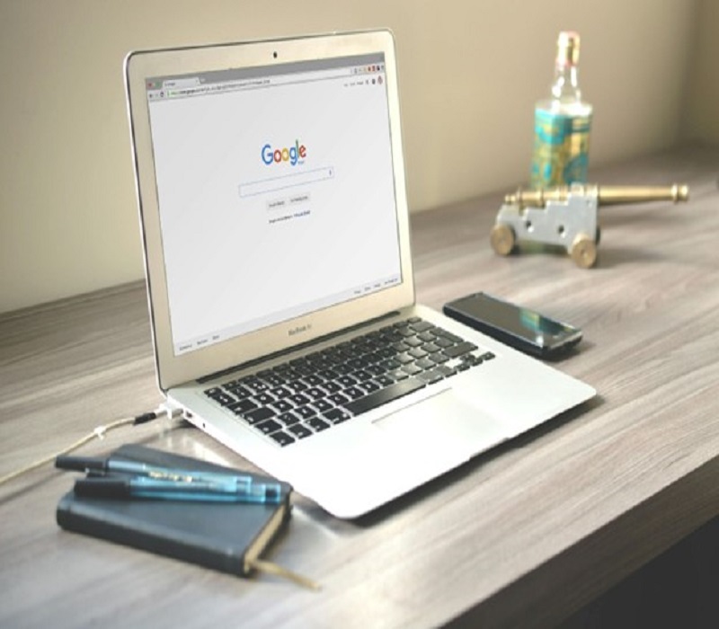 Open laptop on a wooden desk displaying the Google homepage, accompanied by a notebook, pen, smartphone, and decorative items.