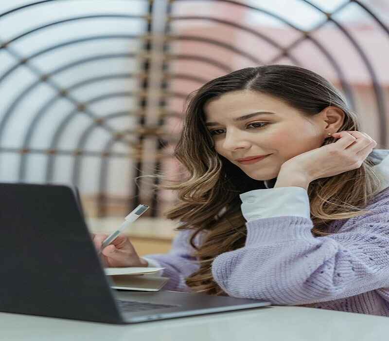 A woman with long hair wearing a lavender sweater sits at a table, holding a pen and looking at her laptop screen thoughtfully, with an arched decorative structure in the background.